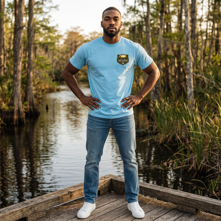 A man wearing a sky blue Fort Denaud Beard and Skin cotton t-shirt with a chest badge graphic, standing on a dock.