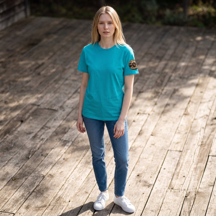Woman wearing a turquoise Fort Denaud Beard and Skin cotton t-shirt with a logo on the sleeve, standing on a wooden deck.